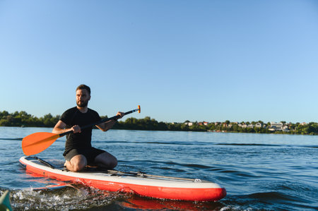 Young sportsman is paddling with orange paddle while kneeling on a sup board, enjoying a summer day...