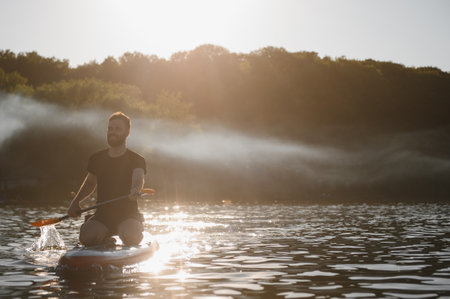 Young man paddling on a sup board at sunset on a lake with light fog, enjoying water sports and...
