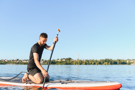 Young man kneeling on a sup board, paddling across a calm lake under the bright sun on a warm summer...