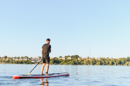 Rear view of sportsman paddling on a sup board on a calm lake in a cityscape, enjoying water sport...