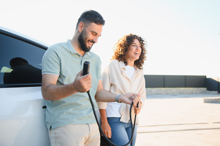 Smiling couple holding charging cable, connecting electric vehicle to power supply at their house, promoting eco friendly transportationの写真素材
