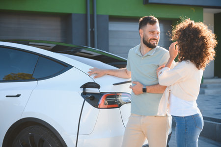 Happy couple stands next to their electric car, which is charging at their home, demonstrating their commitment to green energy and sustainable livingの写真素材