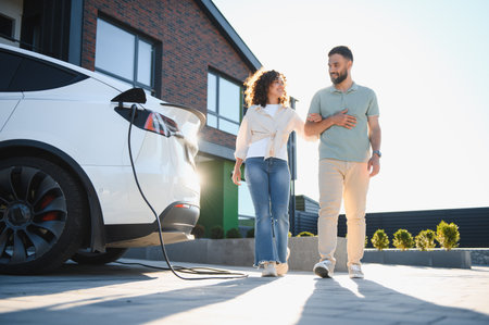 Happy couple walking near their electric car charging in front of their modern house, enjoying sustainable lifestyleの写真素材