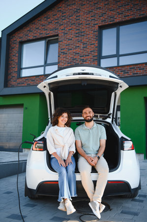 Happy couple sitting in open trunk of electric car, charging at home, enjoying sustainable lifestyleの写真素材