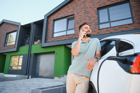 Homeowner charging his electric vehicle and making a phone call in front of his modern house