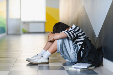 Sad schoolboy sitting alone on the school hallway floor, crying and experiencing the deep pain of bullying and isolationの写真素材