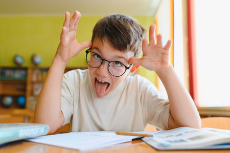 Schoolboy making a funny face and sticking out his tongue while sitting at his desk in a classroom