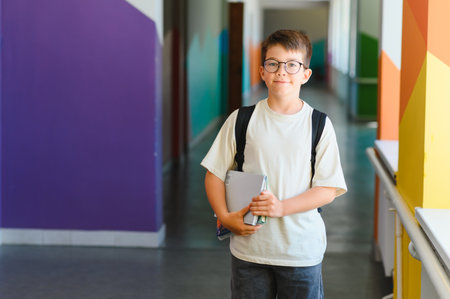 Portrait of a cheerful boy with backpack and books standing in a vibrant school corridor, ready for learningの写真素材