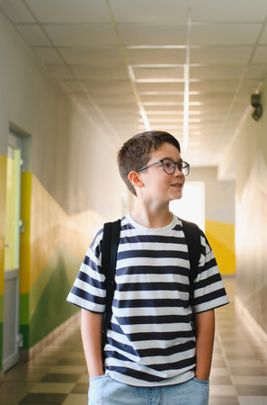 Elementary school student wearing glasses and backpack walking in school hallway, smiling and looking awayの写真素材