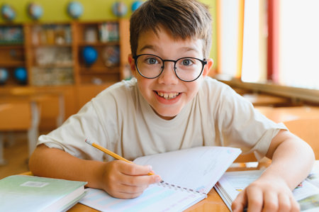 Happy elementary school student writing in a notebook while studying diligently at a classroom desk, showing a cheerful smile and focusの写真素材