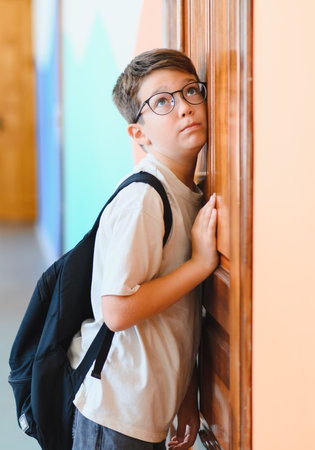 Boy with backpack and eyeglasses carefully listening at the classroom door, trying to eavesdrop on the lessonの写真素材