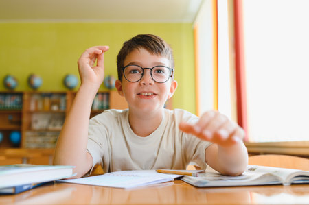 Smart boy with eyeglasses raising hand and actively participating in elementary school lesson, eager to answer a questionの写真素材