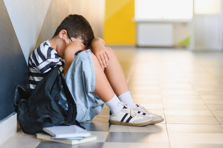 Sad schoolboy sitting alone on the floor in a school corridor, crying and hiding his face, victim of bullyingの写真素材