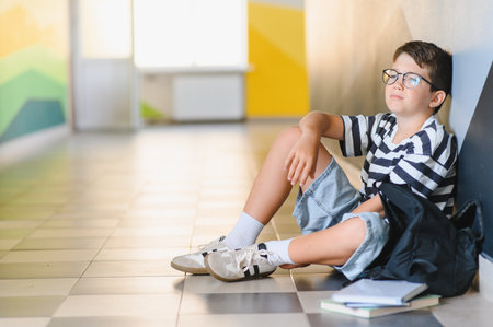 Upset schoolboy sitting on the floor in school corridor with backpack and books, suffering from bullyingの写真素材