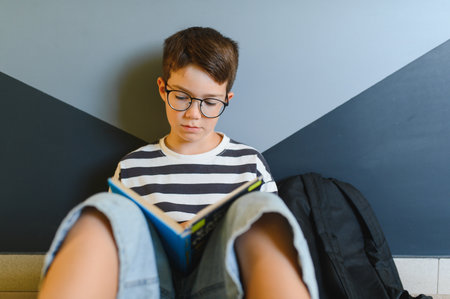Young student reading a textbook while sitting on the floor at school, backpack beside him, focused on his studies and educationの写真素材