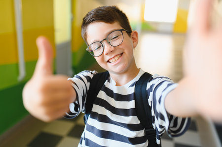 Happy boy with glasses and a backpack giving a thumbs up while taking a selfie in a bustling school hallway, radiating joy and excitementの写真素材