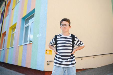 Young student wearing glasses and backpack standing with hands on hips in front of a vibrant multi colored school, ready for learningの写真素材