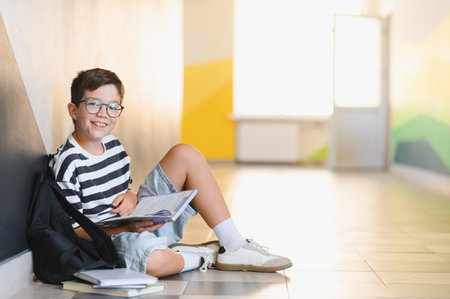 Happy schoolboy sitting on the floor, engrossed in reading a textbook while enjoying a moment of leisure in the school corridorの写真素材