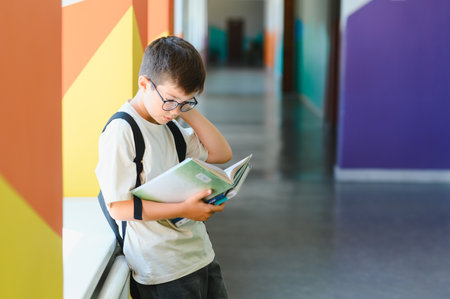 Schoolboy with backpack reading a book in colorful school hallway, concentrating on studying and...