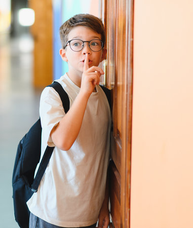 Schoolboy with a backpack making a silence gesture while curiously listening at the classroom door,...