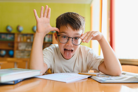 Young boy wearing eyeglasses making a funny face and sticking out his tongue while sitting at a desk...