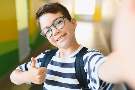 Happy boy with backpack and glasses taking a selfie and gesturing thumbs up in a school hallwayの写真素材