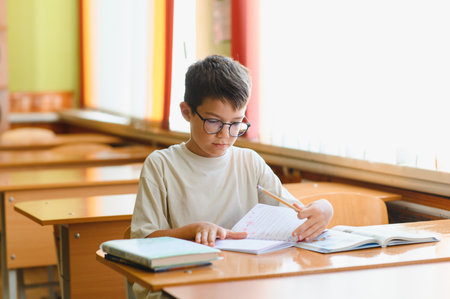 Schoolboy wearing eyeglasses concentrating on studying, writing in notebook at desk in classroom,...