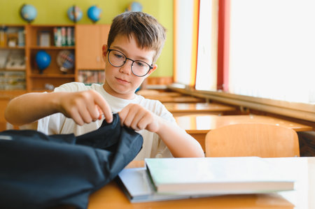 Schoolboy sitting at a desk in a classroom, searching through his backpack for books and school supplies, focused on his studiesの写真素材
