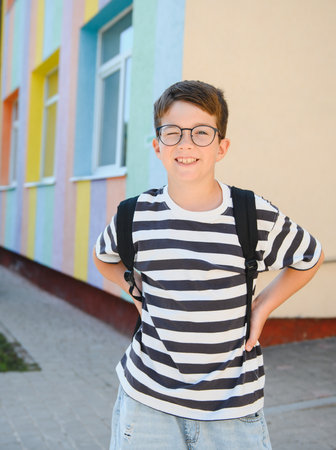 Happy boy winking and smiling while standing near a colorful school, enjoying the excitement of back...