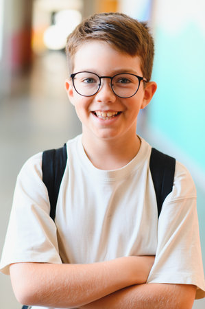 Portrait of a cheerful student wearing glasses and backpack, standing with arms crossed in a school...