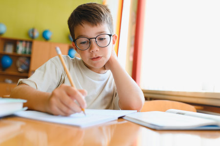 Young boy wearing glasses diligently working on school assignment, writing in notebook at desk in...