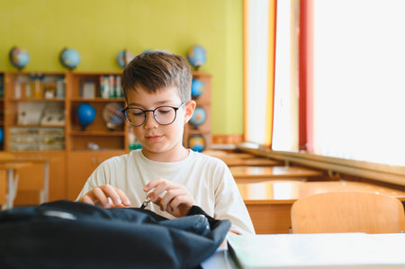Young boy with glasses eagerly opening his backpack at a school desk, preparing for an exciting day of learning and studyingの写真素材