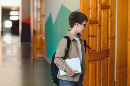 Young student entering the classroom, holding books and wearing a backpack, ready for the school dayの写真素材