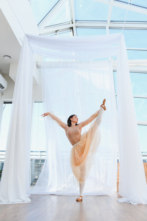 Young ballerina practicing a vertical leg split in a bright dance studio with white curtains and large windowsの写真素材