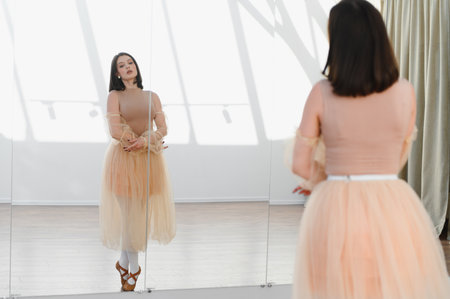 Young female ballet dancer perfecting her poses in a bright dance studio, using a mirror for self...