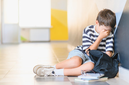 Sad schoolboy sitting on the floor in the school hallway and crying after suffering from bullying by classmatesの写真素材
