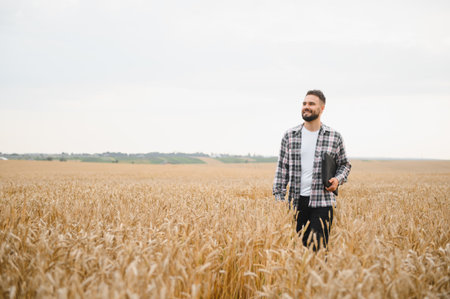 Young farmer walking through golden wheat field, holding tablet and smiling, checking his crops