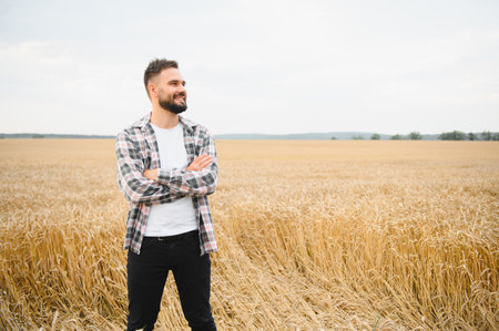 Confident farmer standing with arms crossed in a golden wheat field, smiling brightly while gazing...