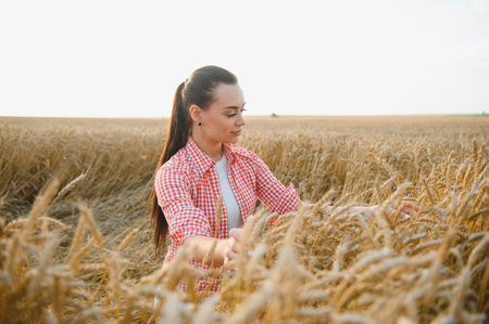 Young woman farmer touching ripe wheat ears in cultivated agricultural field at golden hour,...