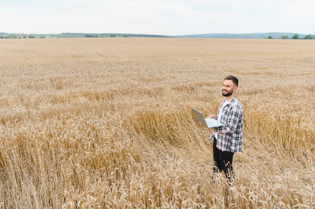 Farmer smiling and using laptop in golden wheat field, implementing modern technology for precision...