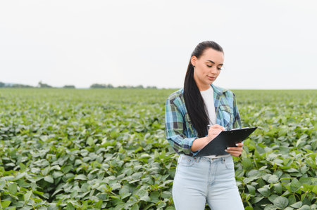 Young woman farmer writing notes on a clipboard while closely inspecting crops in a vibrant soybean...