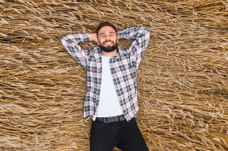 Young farmer lying down and resting with hands behind head in a golden wheat field after a...