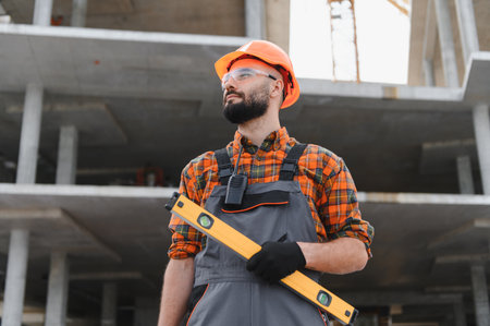 Experienced construction worker wearing a hard hat and safety glasses, holding a spirit level,...