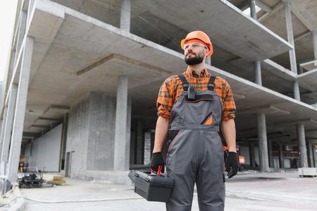 Focused male construction worker in uniform and hard hat holding a toolbox, observing a concrete building under constructionの写真素材