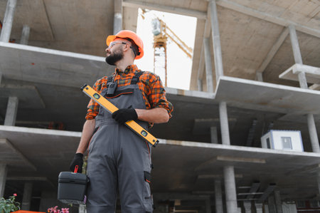 Construction worker wearing hard hat and safety glasses, holding a spirit level and toolbox on a...