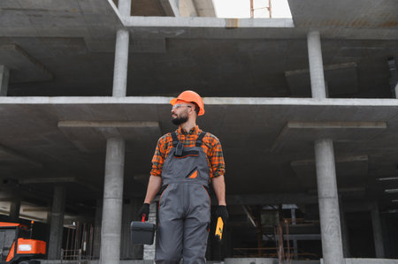 Worker on a construction site wearing safety gear, holding a toolbox and a level, overseeing the building progressの写真素材