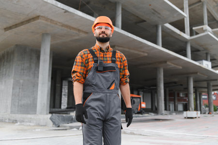 Confident construction worker wearing a hard hat, safety glasses, and overalls, standing at an...