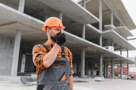 Builder or foreman wearing hard hat and safety glasses, communicating with walkie talkie at an unfinished construction siteの写真素材