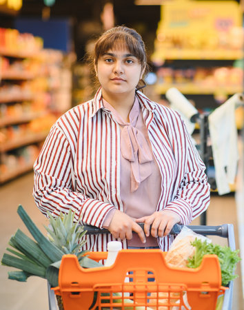 Confident young Indian woman pushing a shopping cart filled with fresh groceries while navigating the bustling aisles of a supermarketの写真素材