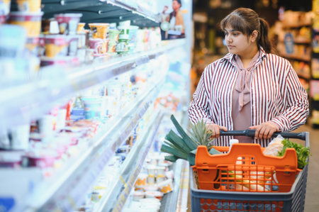 Young Indian woman pushing shopping cart and choosing food products while walking along refrigerated section in grocery storeの写真素材